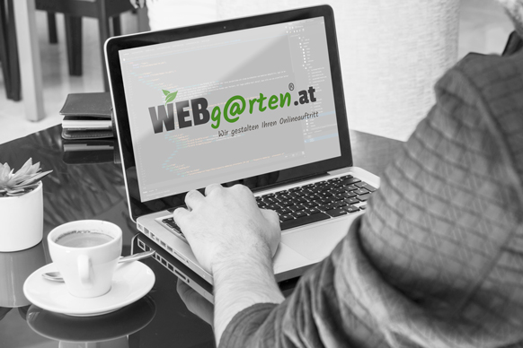 WEBgarten - Webdesign - Braunau | Ein Webentwickler arbeitet an einem Laptop, auf dessen Bildschirm das Logo von WEBgarten.at mit dem Slogan „Wir gestalten Ihren Onlineauftritt“ über einem Programmiercode zu sehen ist; daneben steht eine Tasse Kaffee auf einem Schreibtisch.